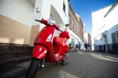 Rote Vespa-Scooter steht auf einem Gehweg in einer Stadtstraße mit Gebäuden im Hintergrund.